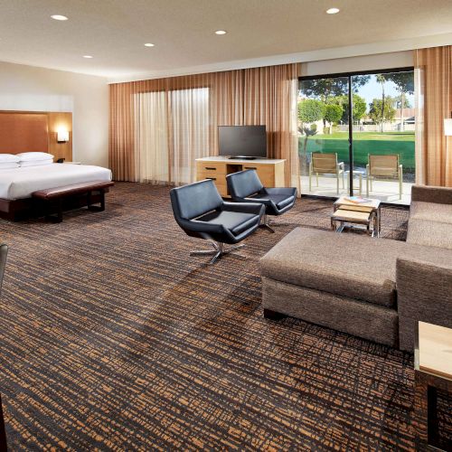 A spacious hotel room with a bed, seating area, TV, large window with a view of greenery, and modern decor.