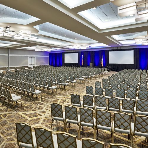 Large conference room with rows of empty chairs facing two projection screens, blue curtains, and overhead lights.