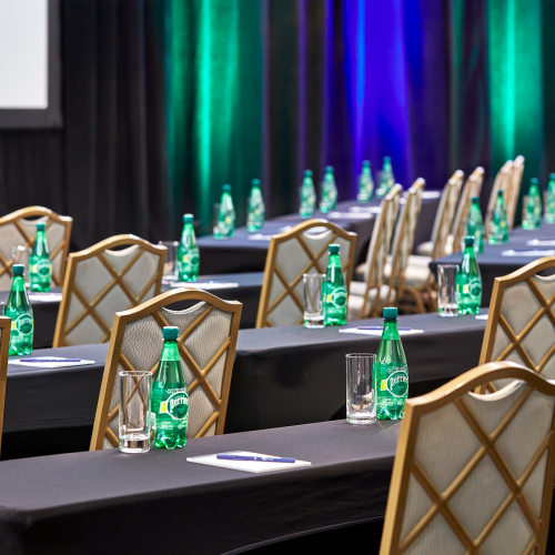 A conference room with rows of tables and chairs, bottled water, glasses, and notepads are neatly arranged on tables.