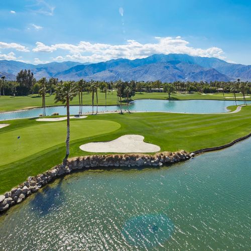A scenic golf course features lush greenery, sand traps, and water hazards set against a backdrop of distant mountains under a clear sky.