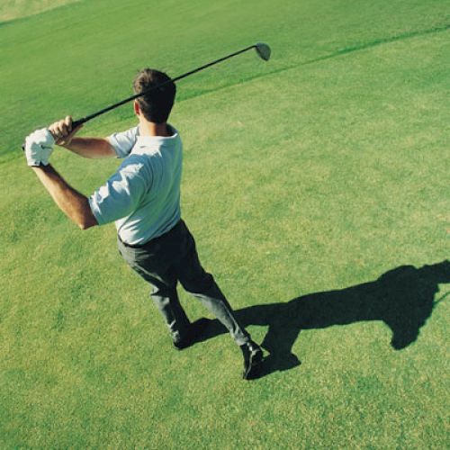 A person is on a golf course completing a swing with a golf club, casting a shadow on the grass.