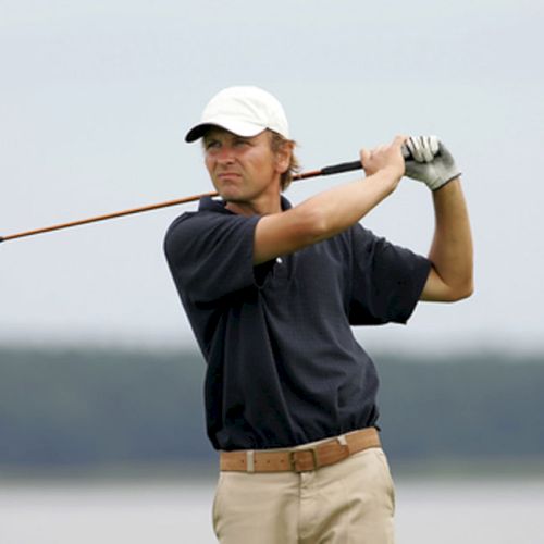 A person in a white cap and gloves is swinging a golf club outdoors, with a blurred background of trees and sky.