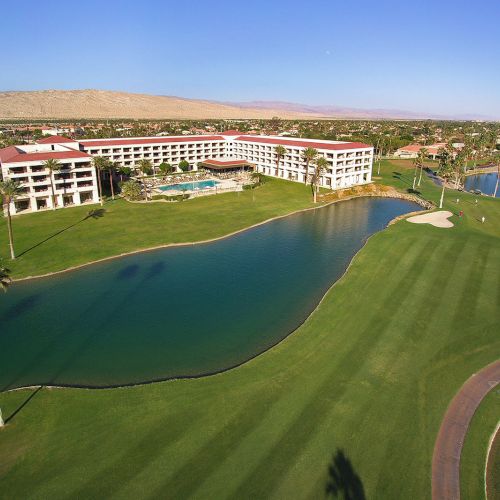 Aerial view of a resort with a pool, palm trees, and a surrounding golf course, set in a scenic landscape.