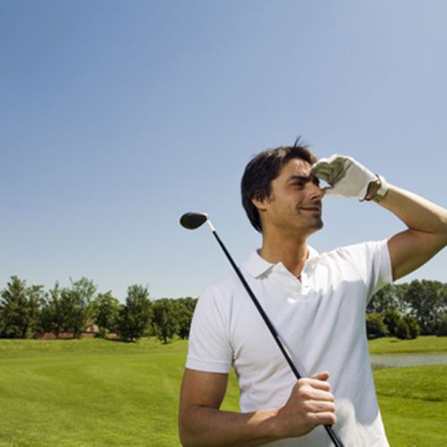 A person in a white shirt on a golf course, holding a golf club and shielding their eyes from the sun.