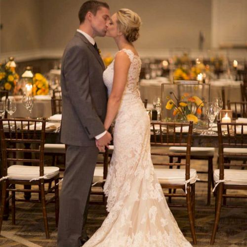 A couple is kissing in a wedding scene, with the bride wearing a lace gown and the groom in a suit, surrounded by elegantly set tables.