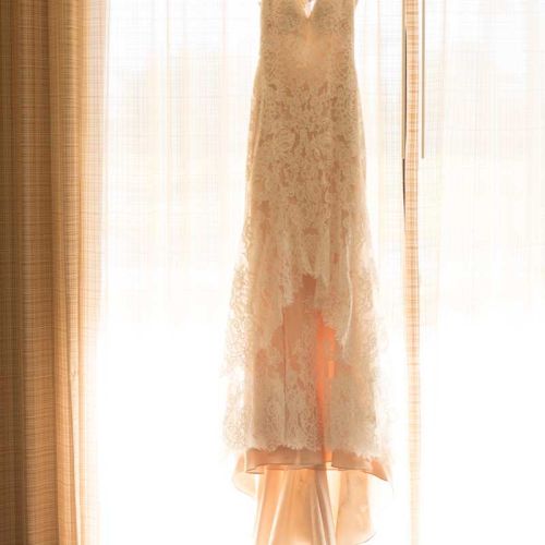 A lace wedding dress is hanging on a wooden hanger in front of a softly lit window.