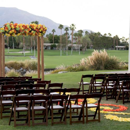 An outdoor wedding setup with chairs and a floral arch on a green lawn, with mountains and palm trees in the background.