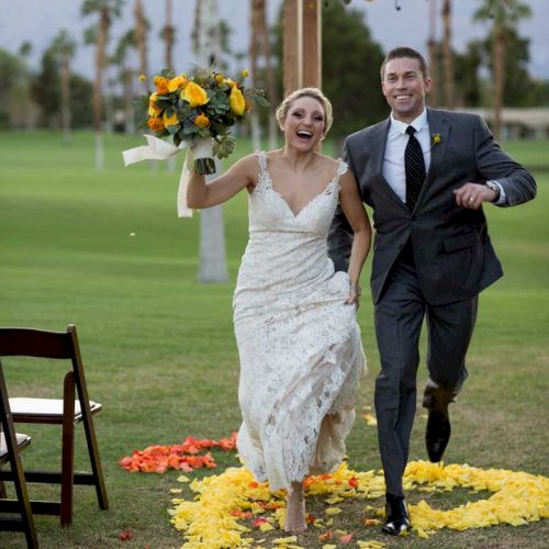 A joyful couple runs down a colorful petal path, the bride holding a vibrant bouquet under a floral arch outdoors.