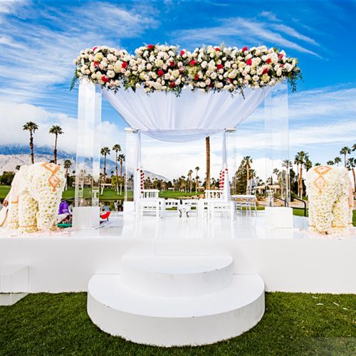 An elegant outdoor wedding setup with a white canopy adorned with flowers, on a lush lawn surrounded by palm trees and a scenic backdrop.