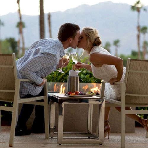 A couple kissing, holding champagne flutes, seated outdoors by a table with a fire, against a backdrop of palm trees and mountains.