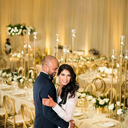 A couple dressed in formal attire smiles in an elegant, decorated banquet hall with white and gold decor, tall candles, and floral arrangements.