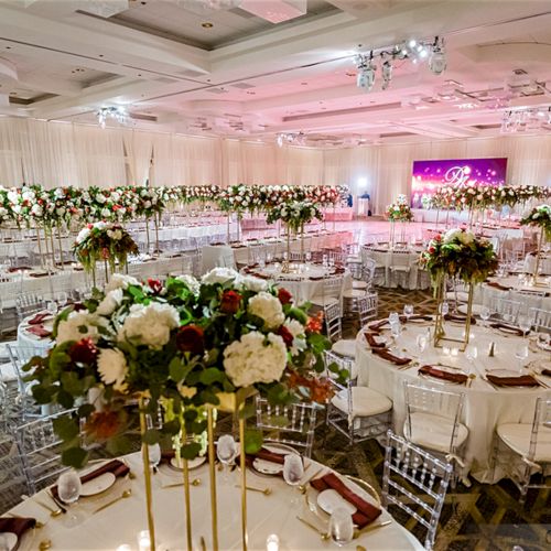Elegant banquet hall set for an event with round tables, elegant centerpieces, and clear chairs under high ceilings with soft lighting.