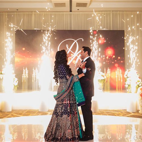 A couple dances on a glossy floor surrounded by flowers and sparklers, with a monogrammed backdrop creating a celebratory atmosphere.