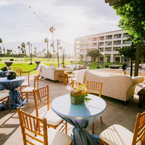Outdoor event setting with tables, chairs, and sofas on a patio. Palm trees, string lights, and a building are visible in the background.