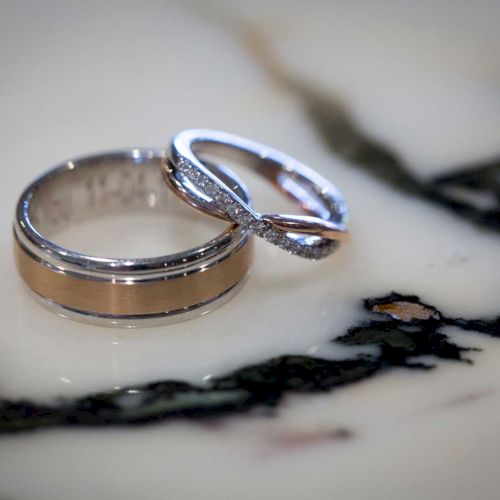 Two intertwined rings, one plain and one with a twisted design and embedded stones, on a marble surface.