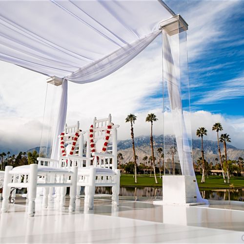 An outdoor wedding setup with white chairs and canopy, red flower garlands, palm trees, and mountains in the background.