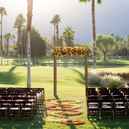 Chairs and a floral arch set up on a sunny lawn for an outdoor event, with palm trees and mountains in the background.