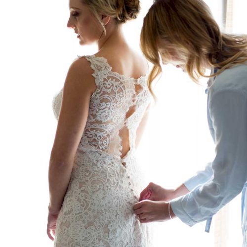 A bride in a lace gown is being helped with her dress by another person in front of a window.