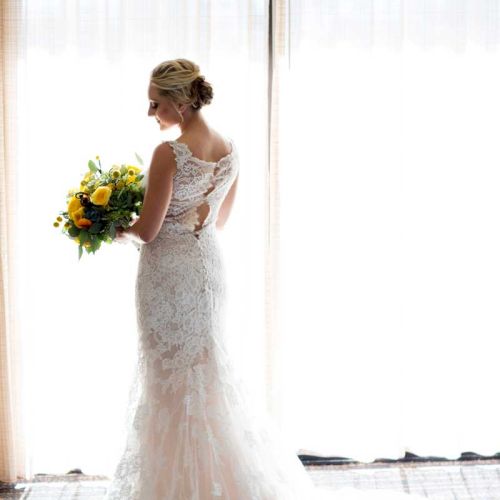 A woman in an elegant white lace wedding gown holds a vibrant bouquet, standing in front of bright curtains.