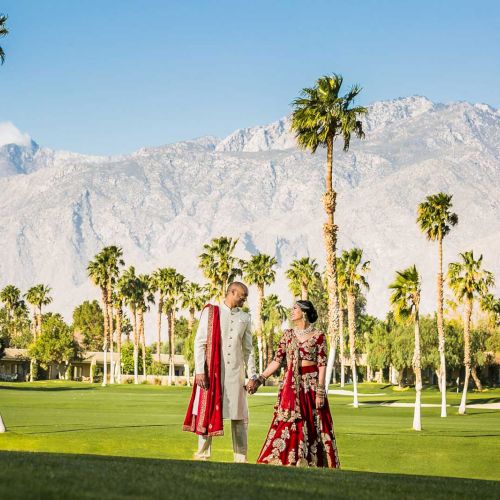 A couple in traditional attire walks hand in hand on a lush lawn, surrounded by palm trees and mountains in the background.