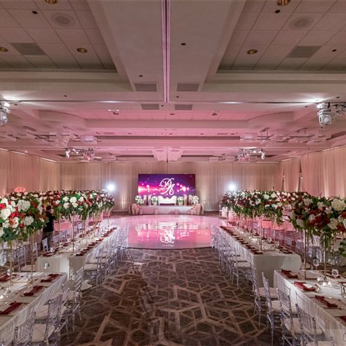 The image shows an elegant, floral-decorated banquet hall with long tables and a lit stage, set for an event or celebration.