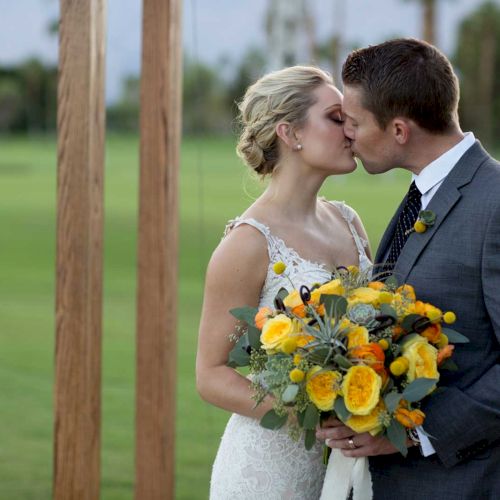 A couple in wedding attire shares a kiss outdoors, with the bride holding a vibrant yellow and orange bouquet.