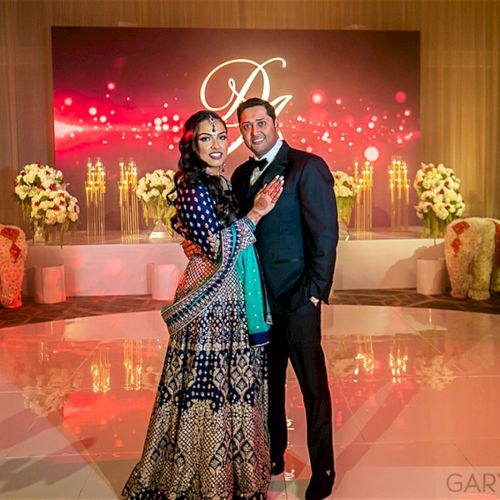 A couple poses in elegant attire at a decorated venue with flowers and candles, against a backdrop of red lights and an emblem.