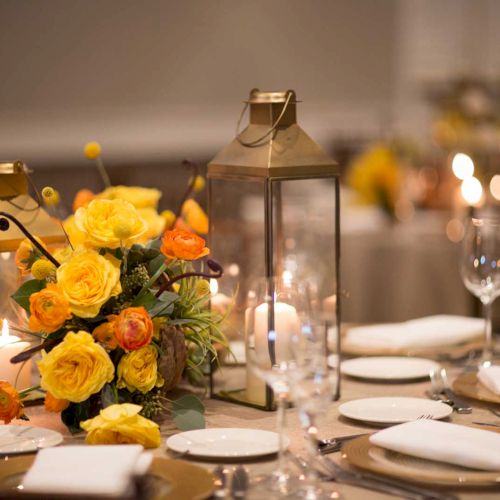 Elegant table setting with glassware, white plates, yellow and orange flower centerpiece, and lanterns, creating a warm ambiance.