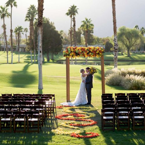 A couple stands under a floral arch on a lush lawn with palm trees, surrounded by empty chairs in a wedding setup.