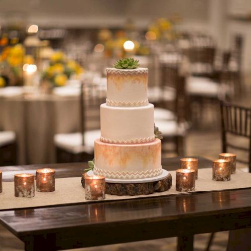 A three-tiered wedding cake with a succulent on top, surrounded by candles on a wooden table, with a decorated venue in the background.