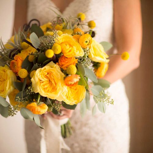 A person is holding a vibrant bouquet with yellow and orange flowers, featuring greenery, in a warm setting.