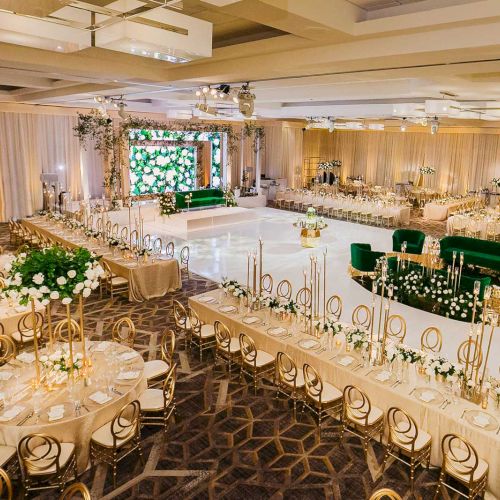 Luxurious event venue with elegant tables, golden chairs, and green accents, set for a sophisticated celebration, featuring floral decor.