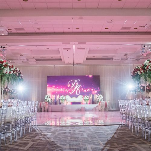 An elegant event venue with floral decorations, transparent chairs, and a stage with an illuminated backdrop featuring a monogram.