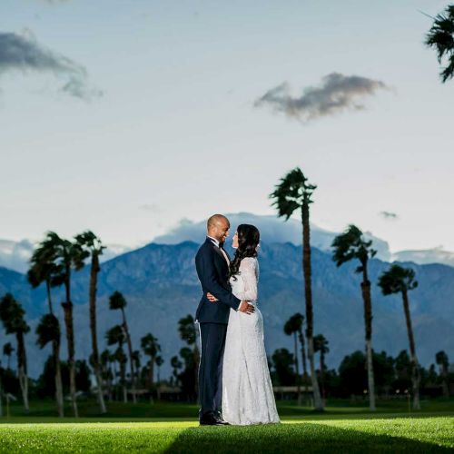 A couple stands embracing on a grassy field with palm trees and mountains in the background under a cloudy sky.