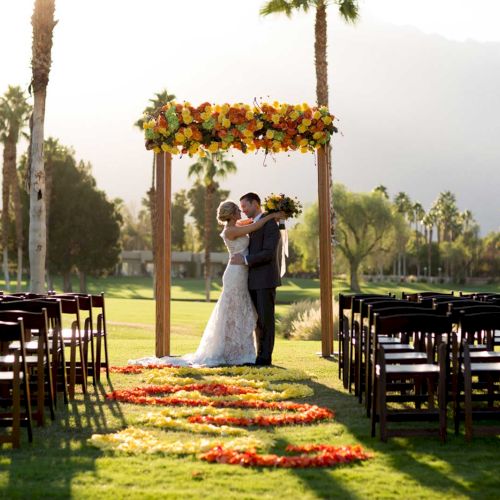 A wedding couple embraces under a floral arch on a sunny outdoor lawn with empty chairs and palm trees in the background.
