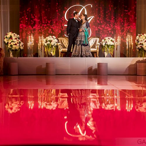 A couple stands on a decorated stage with a red pixel backdrop, floral arrangements, and reflections on the shiny floor.