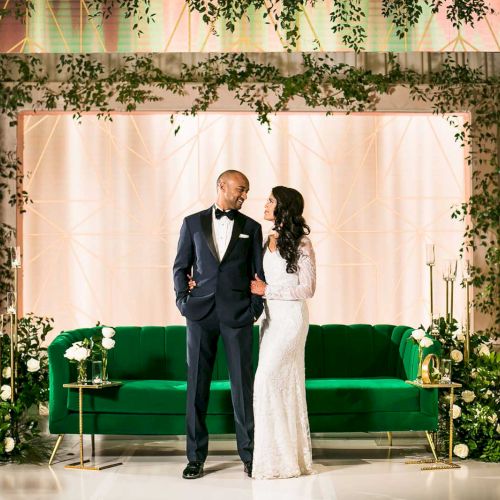 A couple stands on a decorated stage with greenery, a green couch, and elegant lighting, creating a romantic atmosphere.
