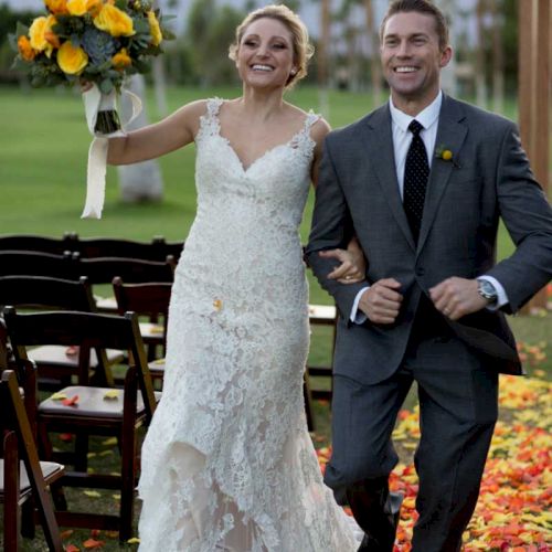 A joyful couple walks arm-in-arm down an aisle covered with petals, with the bride holding a bouquet, outdoors.