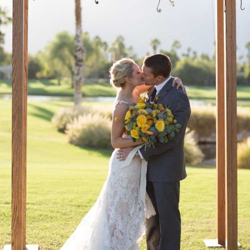 A couple is kissing under a floral arch at an outdoor wedding ceremony, with the bride holding a bouquet.