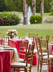 Outdoor event setup with tables covered in red and white cloth, surrounded by chairs, and a floral centerpiece, near a fountain.