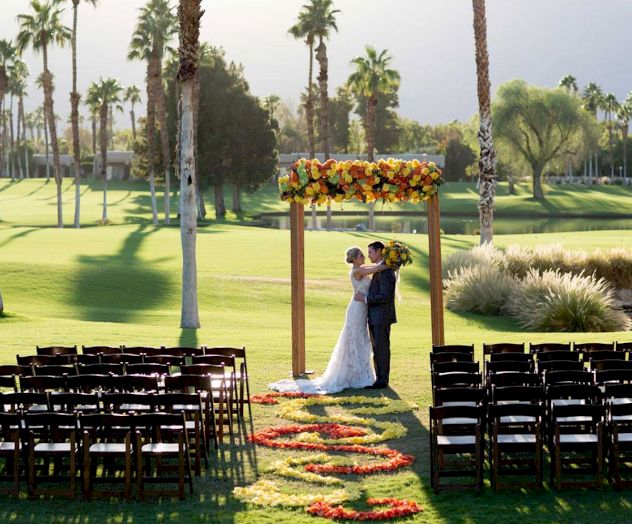 A couple is standing under a floral arch on a grassy field, with decorated chairs and palm trees in the background, suggesting a wedding.