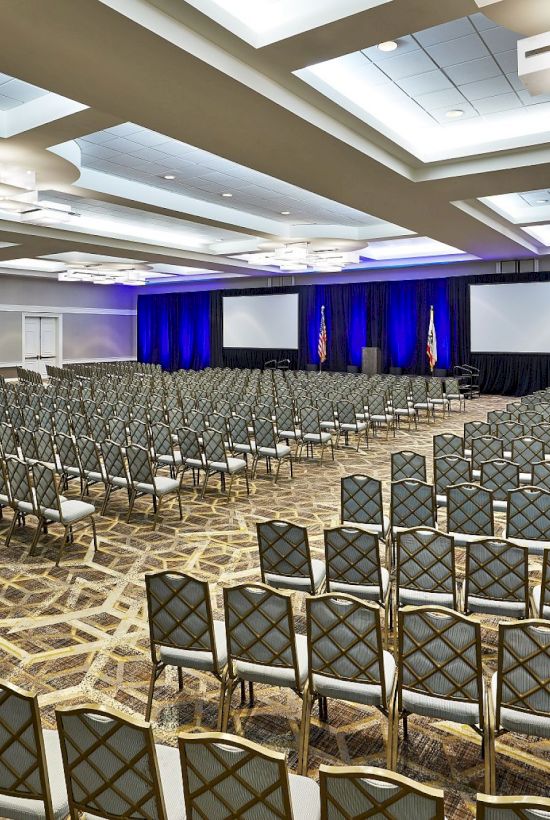 An empty conference room with rows of chairs, two large screens, and blue curtains, ready for a presentation or event.