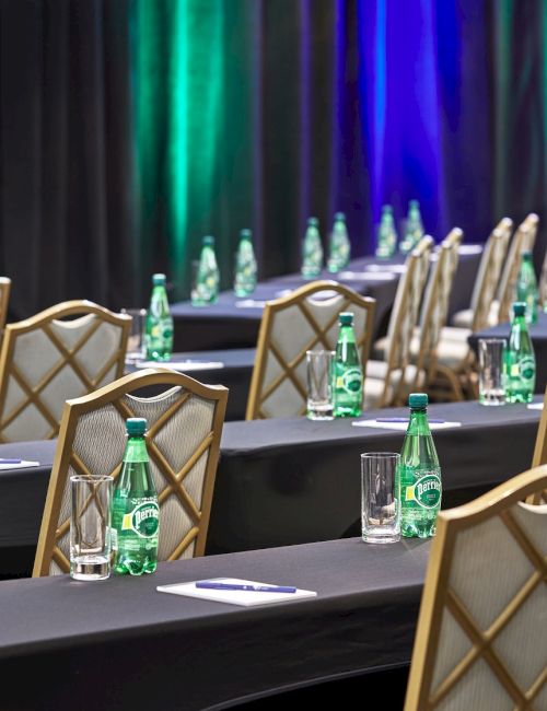 The image shows a conference room setup with neatly arranged tables, chairs, water bottles, glasses, and notepads in a well-lit space.