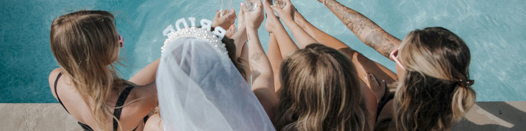 Four women sit poolside in swimsuits, one wearing a veil, touching feet in the water while toasting with drinks, highlighting celebration.