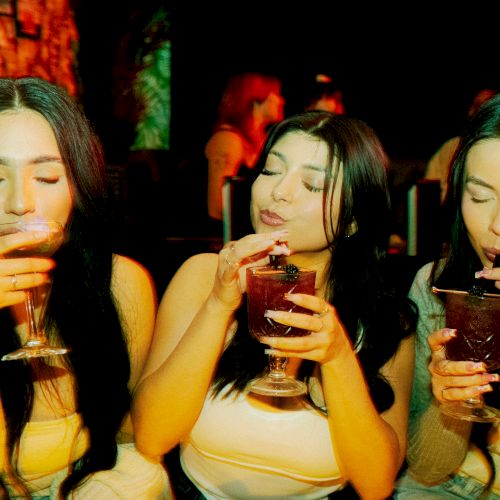 Three people are sitting together, each sipping a drink from a glass, while enjoying what seems to be a lively atmosphere.