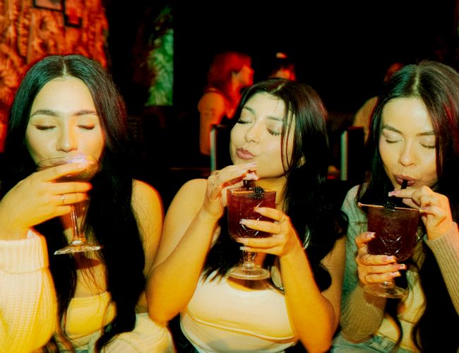 Three people are sitting together, each sipping a drink from a glass, while enjoying what seems to be a lively atmosphere.