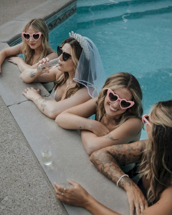 Four people are in a pool wearing sunglasses; one has a bridal veil, suggesting a bachelorette party theme, with drinks nearby.