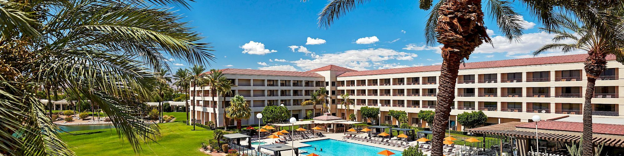 The image shows a hotel with a large pool area, surrounded by palm trees and a grassy lawn under a clear blue sky.