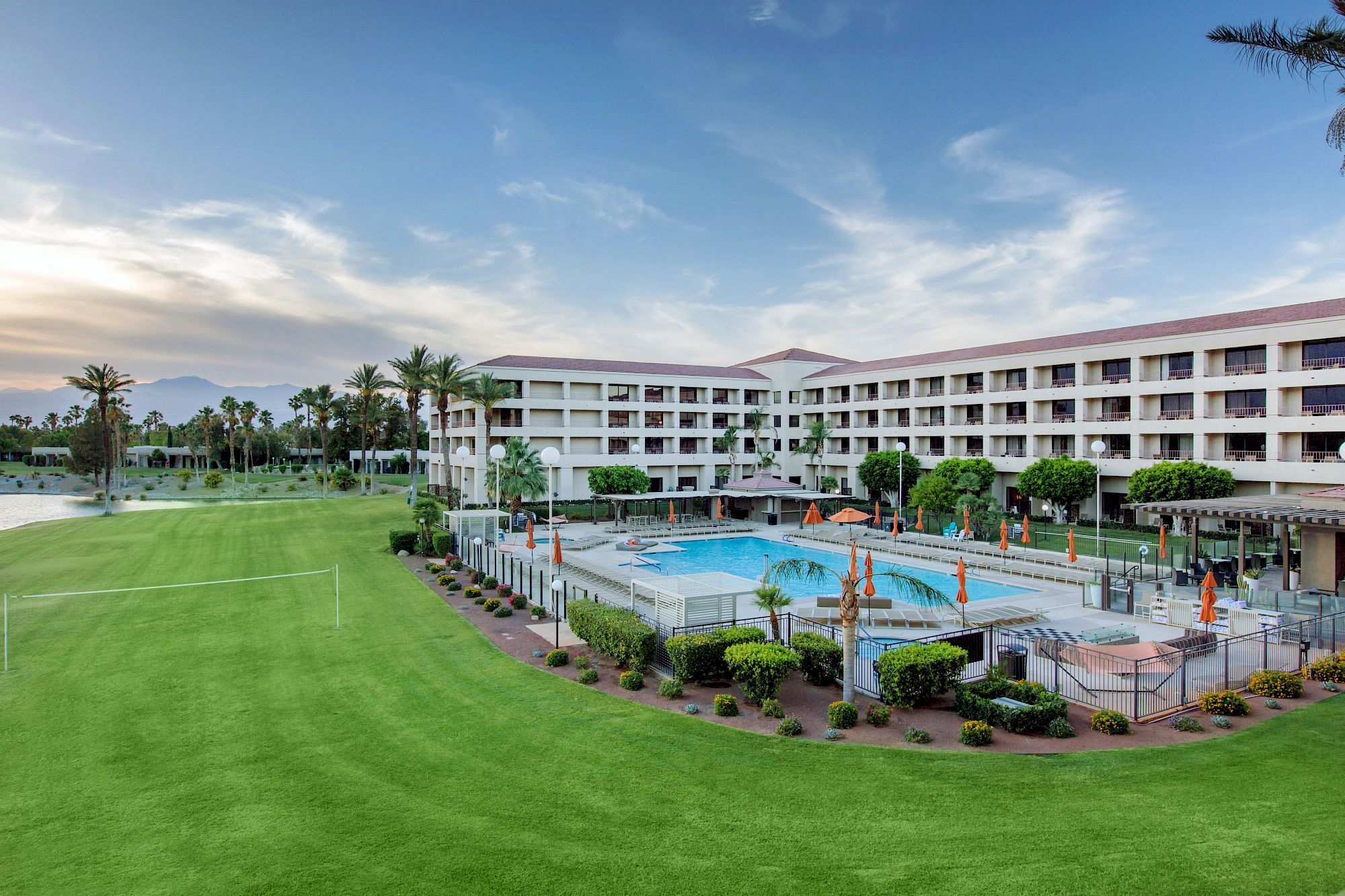 A resort-style hotel with a large green lawn, a pool area, palm trees, and a multi-story building surrounding the courtyard, under a blue sky.