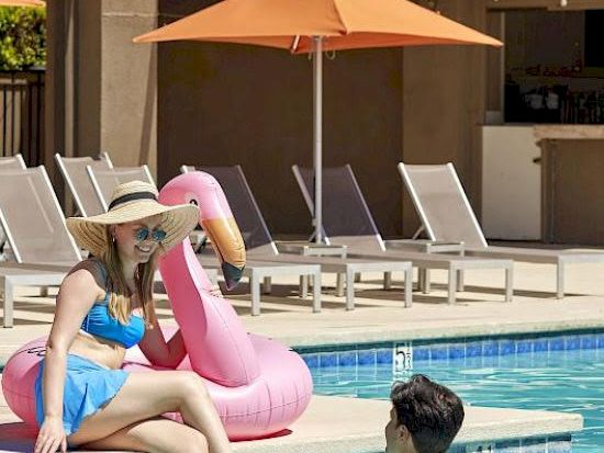 A woman in a blue swimsuit sits by a pool with a pink flamingo float, while a man sits in the shallow end near the edge, sunny resort surroundings. end.